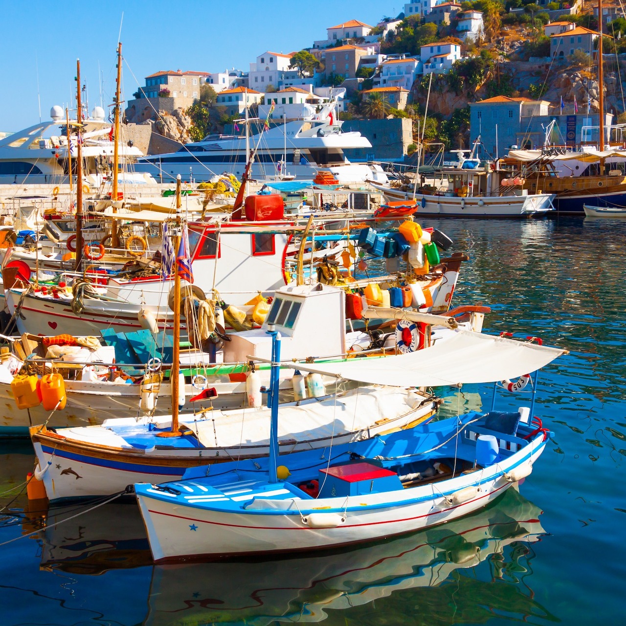 Colorful fishing boats moored in a Greek harbor below white hillside houses.