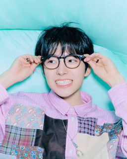 Young man with black hair adjusting round glasses, wearing pastel patchwork jacket, mint background