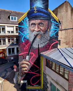 Large mural of an elderly sailor smoking a pipe on the side of a building in a town street