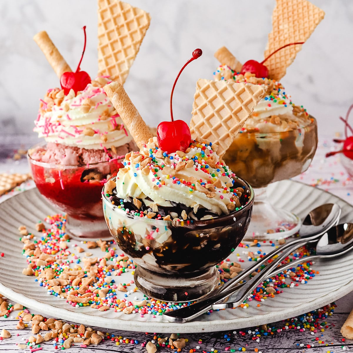 Three ice cream sundaes with waffle cones, whipped cream, cherries and colorful sprinkles in glass bowls