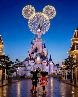 Fireworks forming Mickey Mouse ears above pink Disneyland castle at twilight with two silhouetted visitors