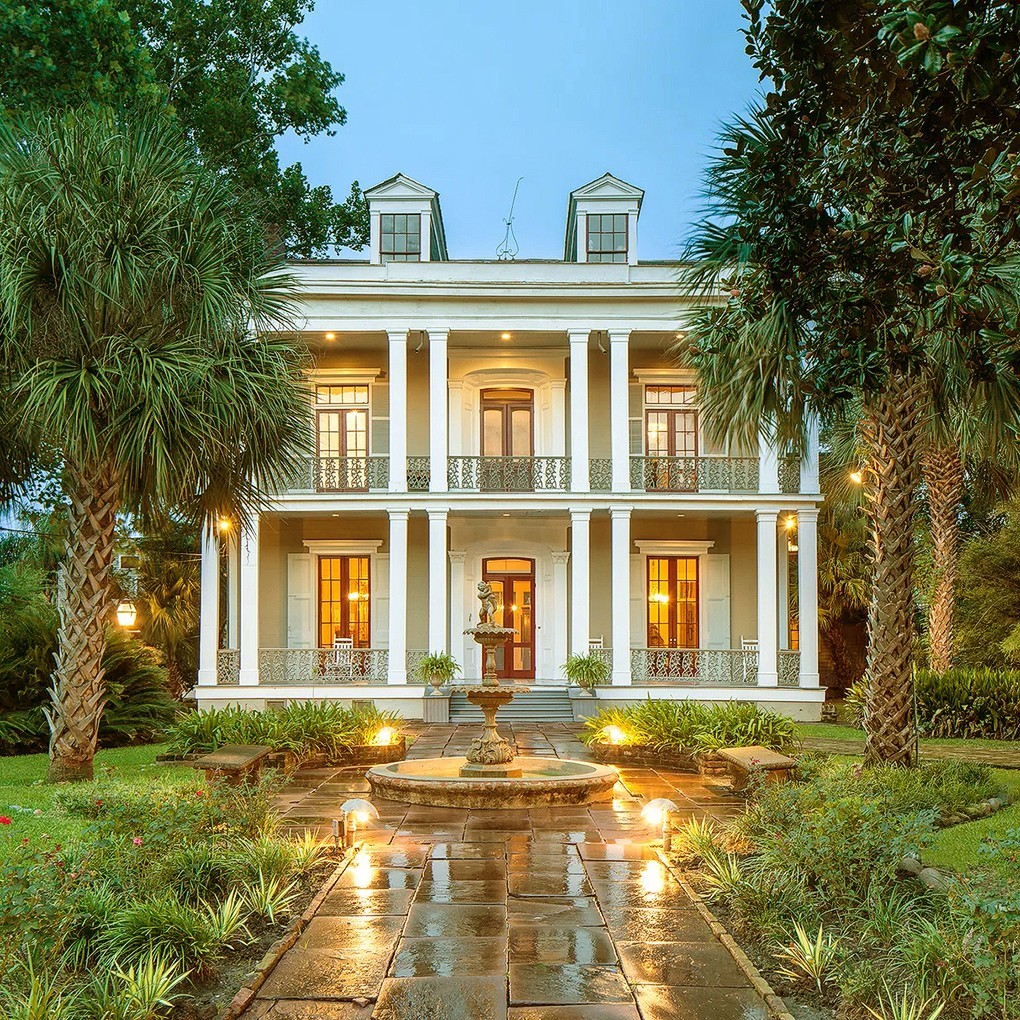 White Greek Revival mansion with tall columns and balconies, lit up at twilight with palm trees and pathway