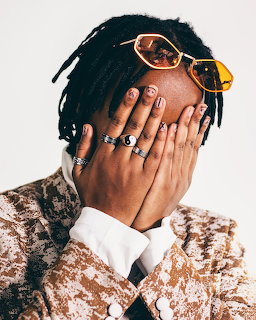 Person covering face with ring-adorned hand, wearing rose gold sunglasses and sequined jacket