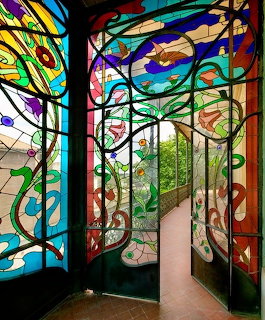 Art Nouveau stained glass interior with colorful floral patterns and curved doorway framing garden view