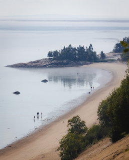 Sandy beach and dunes along calm water with people walking and a forested peninsula at Tadoussac, Quebec