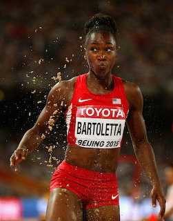 Female athlete in red USA uniform running through water splash at track and field competition