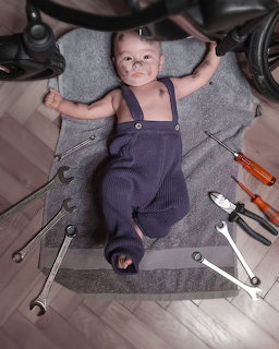 Smiling baby in blue overalls lying on a rug with hand tools around them indoors