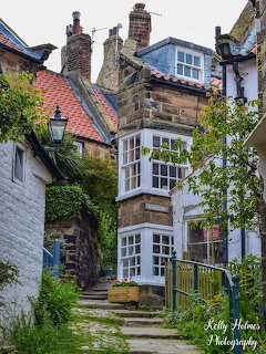 Stone cottage with bay window on a narrow village lane with steps, lamp post, and lush greenery