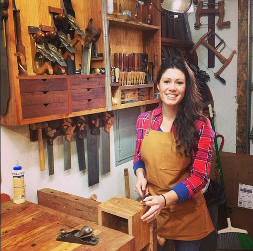 Smiling woman in a woodworking workshop beside a bench with hand tools on the wall