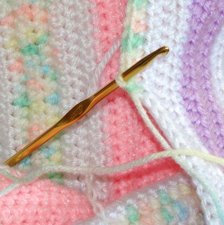 Gold crochet hook on a pastel pink, white, and lavender crocheted baby blanket