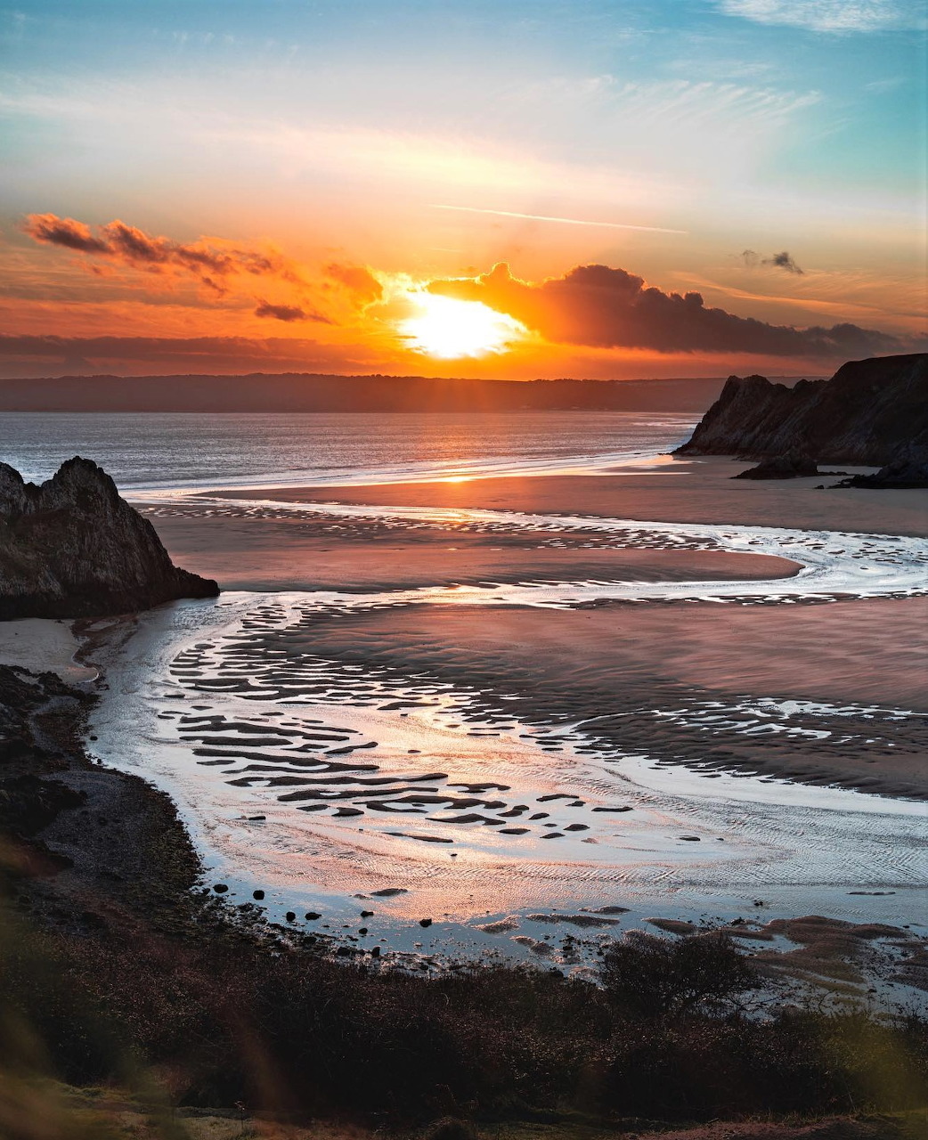 Sunset over a beach with tidal channels, sea, and dark coastal cliffs