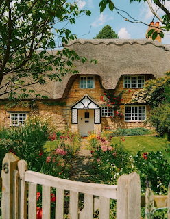Charming English cottage with thatched roof, stone walls, and vibrant flower garden behind white picket fence