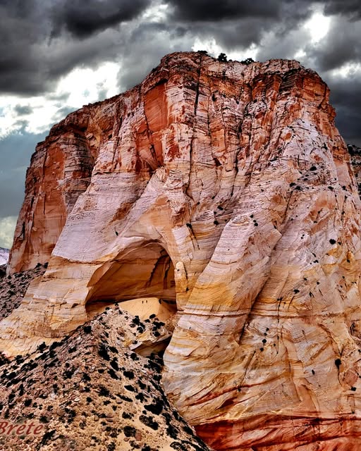 Towering layered sandstone rock formation with arch-like opening under dark stormy sky in Utah