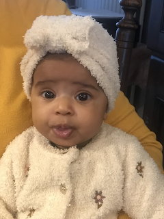 Baby wearing a white towel headwrap and cream outfit, looking at the camera indoors
