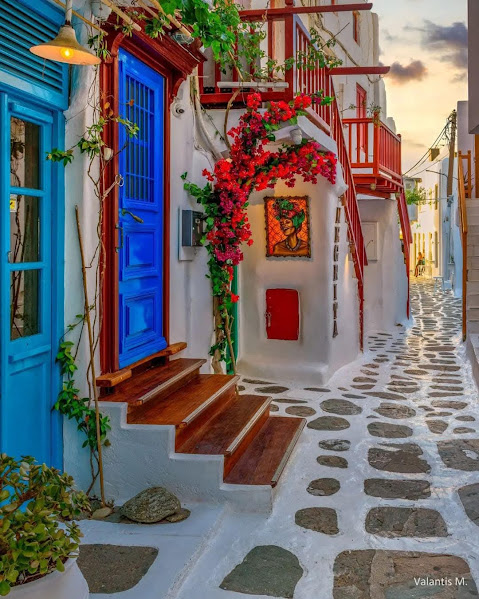Narrow Greek island street with white buildings, blue door, red flowers, and sunset sky