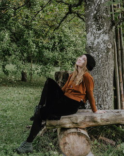 Woman in orange shirt sitting on wooden bench leaning against tree trunk in lush green park
