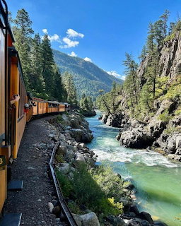 Yellow train on narrow gauge tracks beside turquoise river in a forested mountain gorge