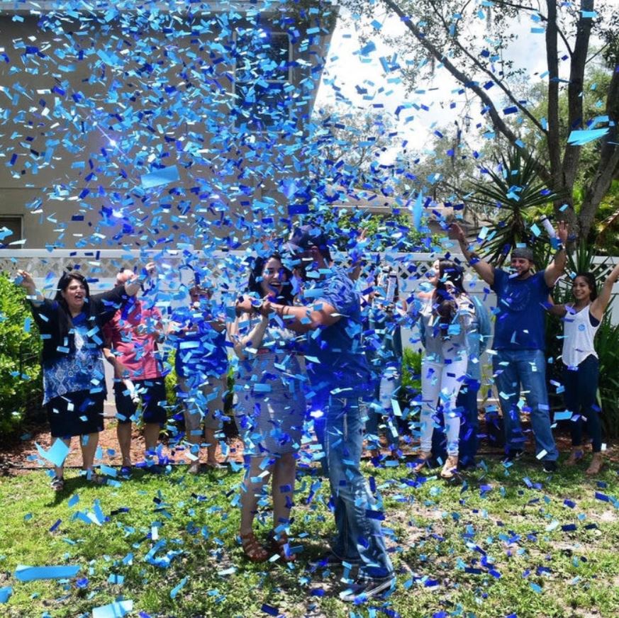 Couple hugging at an outdoor gender reveal with blue confetti and cheering family and friends