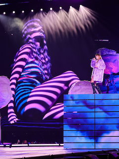 Taylor Swift performing on a concert stage with purple lighting and a large screen backdrop
