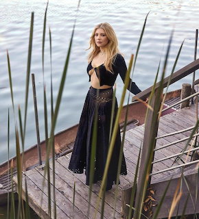 Blonde woman in black outfit standing on a wooden dock by a lake with green reeds in foreground