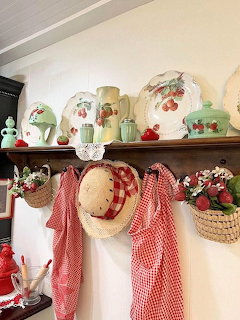 Vintage kitchen shelf with jadeite green dishes, strawberry ceramics, straw hat and red gingham aprons hanging below