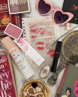 Flatlay of pink beauty products, heart sunglasses, kiss-marked book page, and vintage accessories