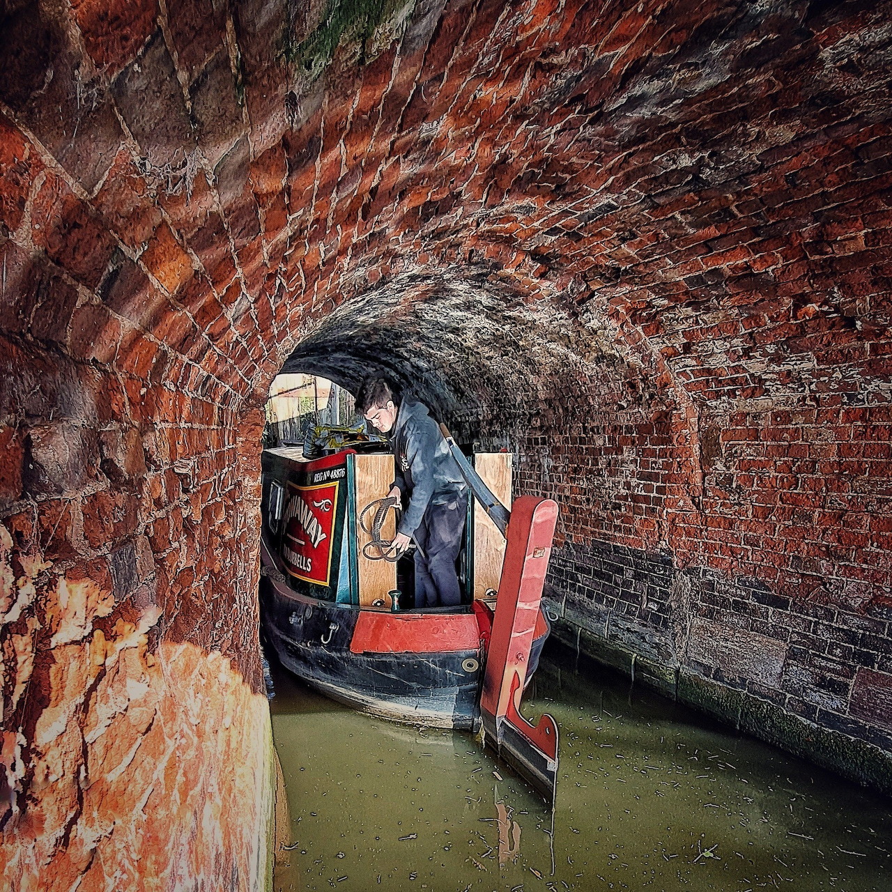 Narrowboat in brick-arched canal tunnel with person standing at stern, murky green water below vaulted brick ceiling