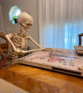 Skeleton seated at a wooden table assembling a jigsaw puzzle indoors