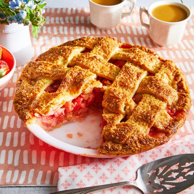Lattice-topped strawberry-rhubarb pie with slice removed, showing red fruit filling, on white plate with checkered cloth