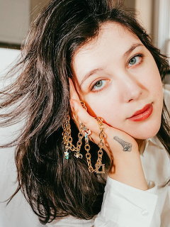 Woman with dark wavy hair and dangling earrings resting hand near face, looking at camera