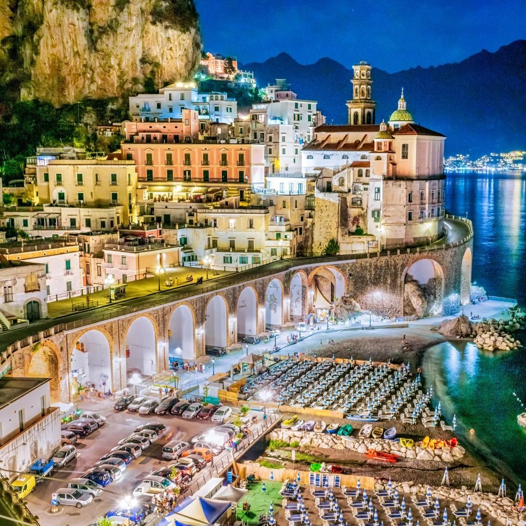 Coastal town at night on the Amalfi Coast with cliffside buildings, arched road, harbor, and sea