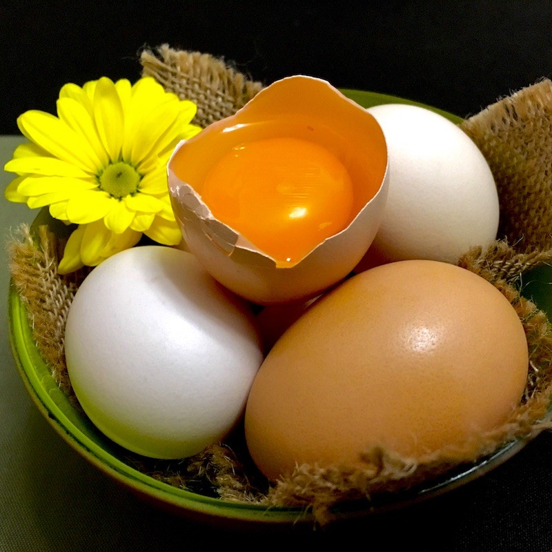 Fresh white and brown chicken eggs in a green bowl with one cracked open showing yolk and a yellow flower