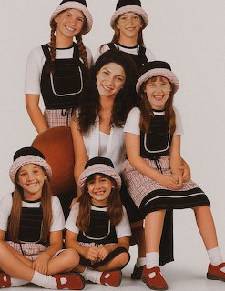 Group of girls and woman wearing matching black pinafore outfits with plaid hats posing together