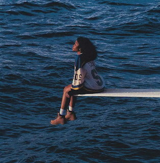 Woman sitting on a diving board over dark blue ocean water, looking up, wearing a sports jersey and boots