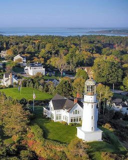 White lighthouse with attached house on grassy grounds, surrounded by fall foliage and coastal scenery