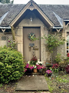 Wooden cottage door with wreath, surrounded by stone walls, slate roof, and vibrant garden flowers on pathway