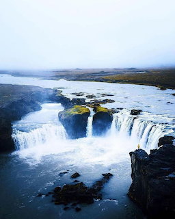 Wide waterfall flowing over dark rocky cliffs into a river with misty landscape in background