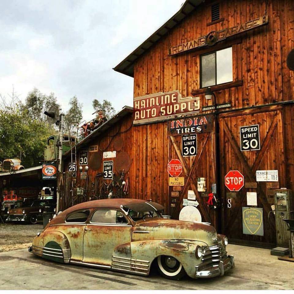 Rusty vintage Chevrolet parked in front of an old wooden auto supply garage with road signs