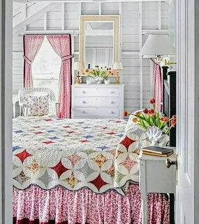 Cozy farmhouse bedroom with colorful patchwork quilt, pink curtains, white walls, and fresh tulips