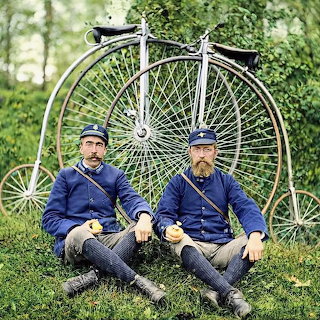 Two uniformed men sitting on grass before penny-farthing bicycles in a colorized vintage photograph.