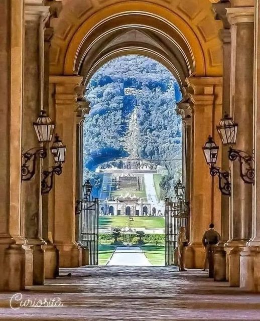 Ornate palace archway with columns and lanterns framing a view of formal gardens and fountains