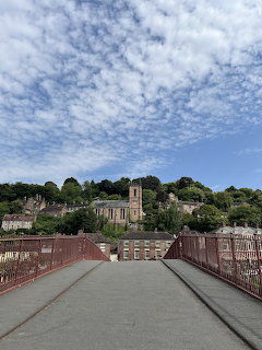 Red iron bridge leading to a hillside town with a church tower under a cloud-filled blue sky