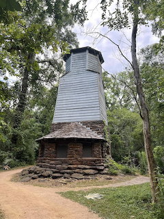 Tall wooden water tower with stone base surrounded by green trees on a dirt path