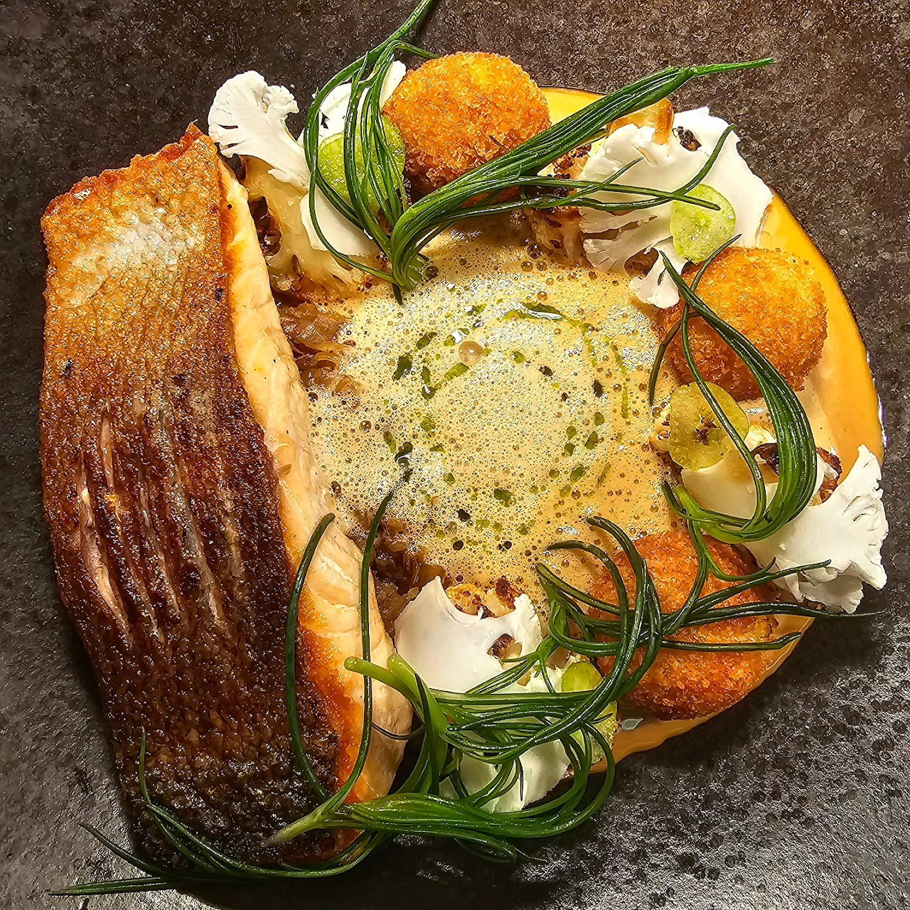 Seared fish fillet with golden skin, green sea vegetables, orange puree, and white flowers on dark plate