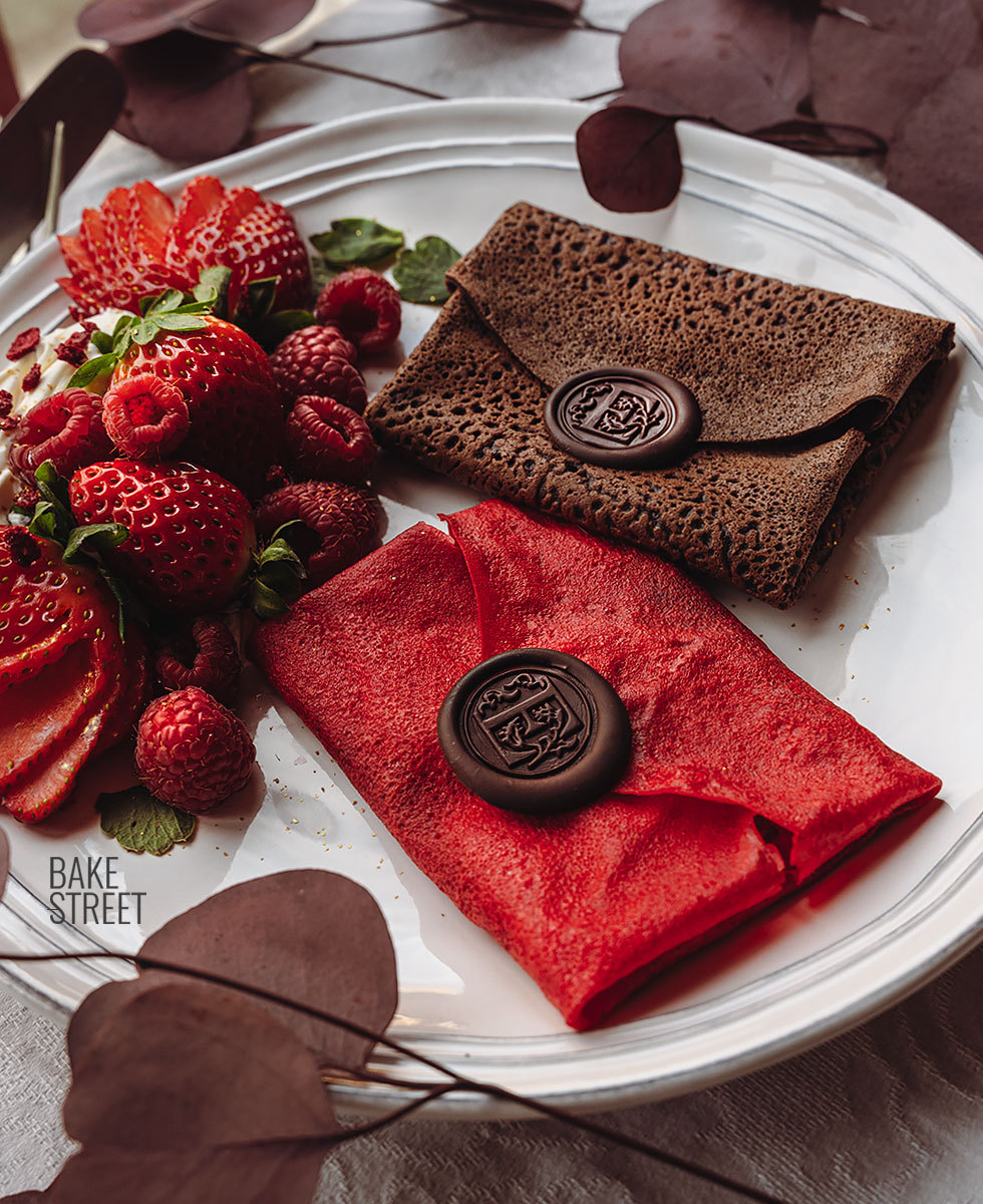 Red and chocolate crepes shaped like envelopes with wax seals, served with strawberries on a white plate