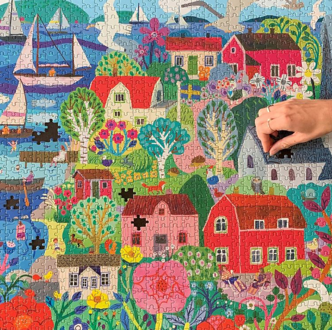 Hand placing a puzzle piece on a colorful illustrated village scene with houses, trees, flowers, and water