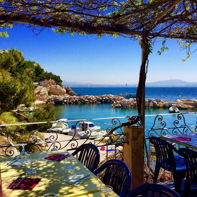 Shaded terrace with tables and chairs overlooking a rocky cove and blue sea