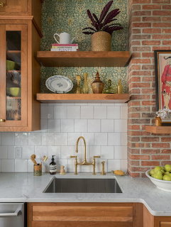 Kitchen corner with brick wall, subway tile, concrete counter, sink, gold faucet, and wooden shelves with plants