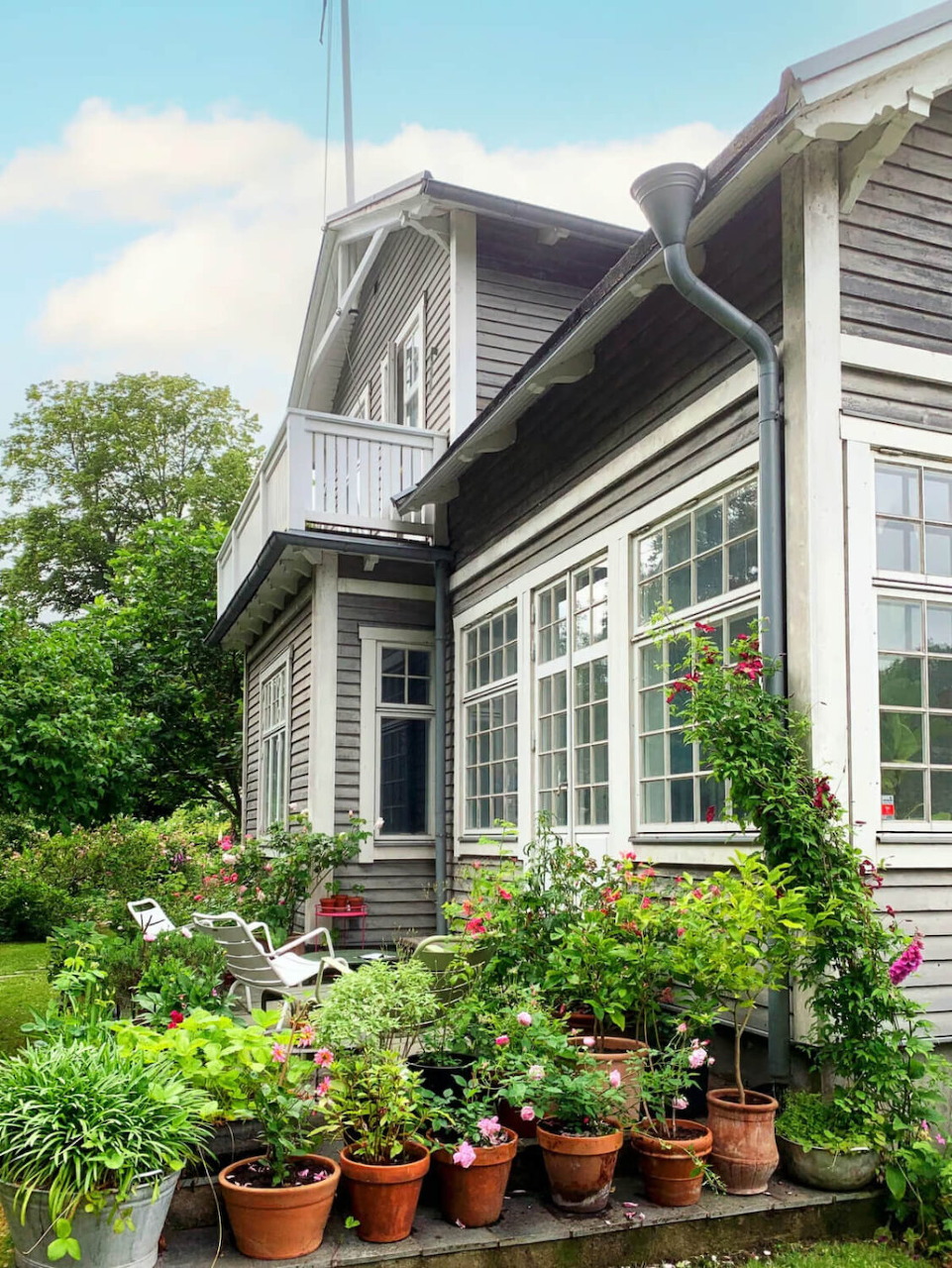 Gray house with white porch and balcony, garden beds with flowers in terracotta pots in foreground