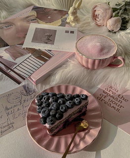Slice of blueberry cake on a pink plate beside a pink drink, roses, and magazines